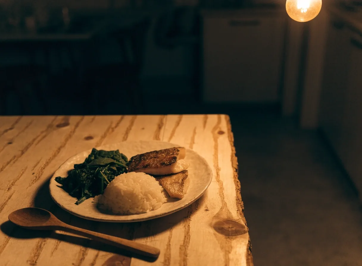 A matte plate of rice, greens and grilled fish on a birch table under warm rim light
